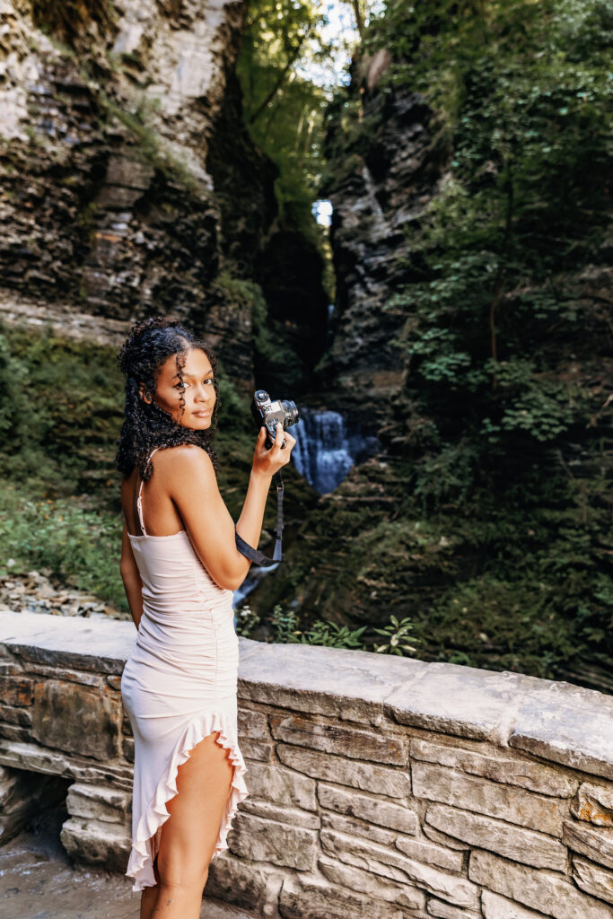 A person holding a camera stands beside a stone wall, with lush greenery and a waterfall in the background, possibly at a nature park.