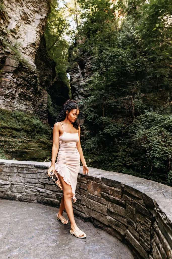A person in a pale dress walks along a stone path surrounded by lush vegetation and rocky cliffs in a natural setting.