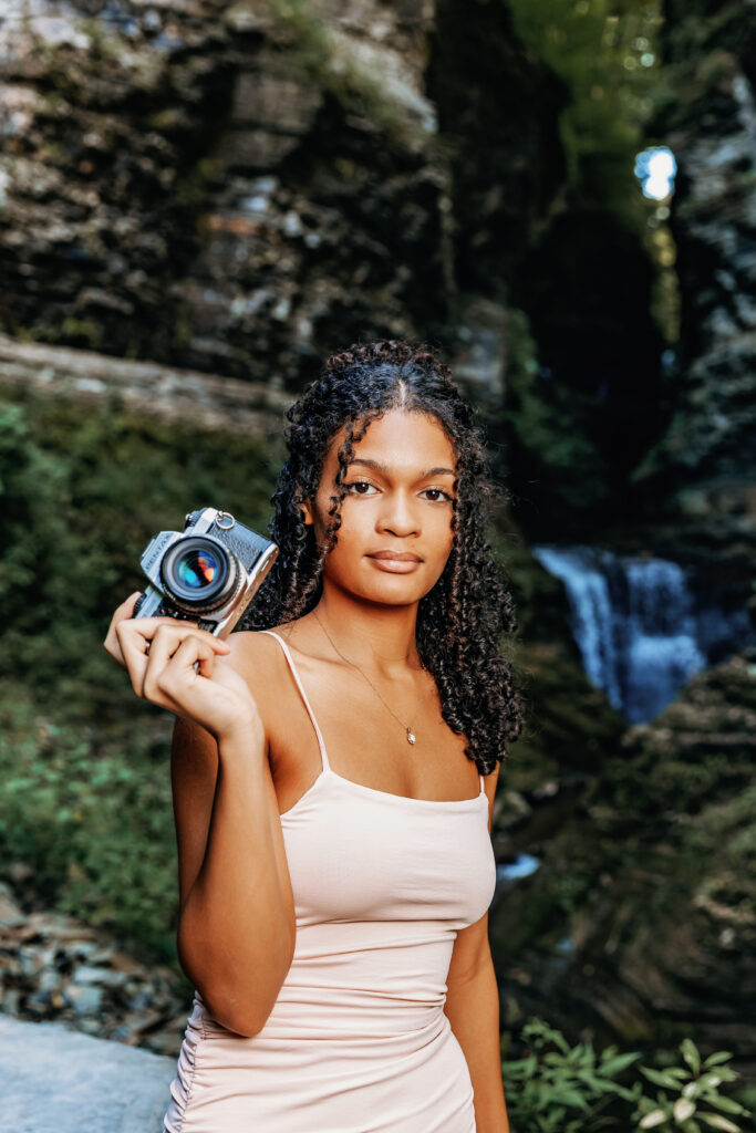 A person holding a camera stands in front of a rocky landscape with a waterfall and greenery in the background.