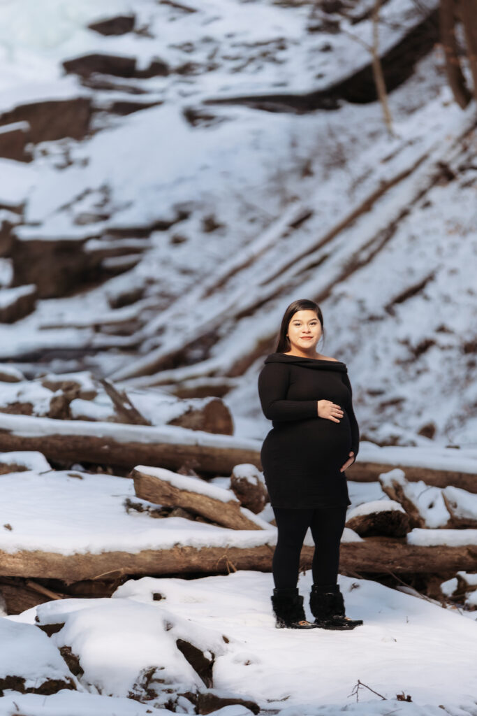 A person stands on snowy ground, surrounded by logs and rocky terrain, wearing a black outfit, in an outdoor winter setting.