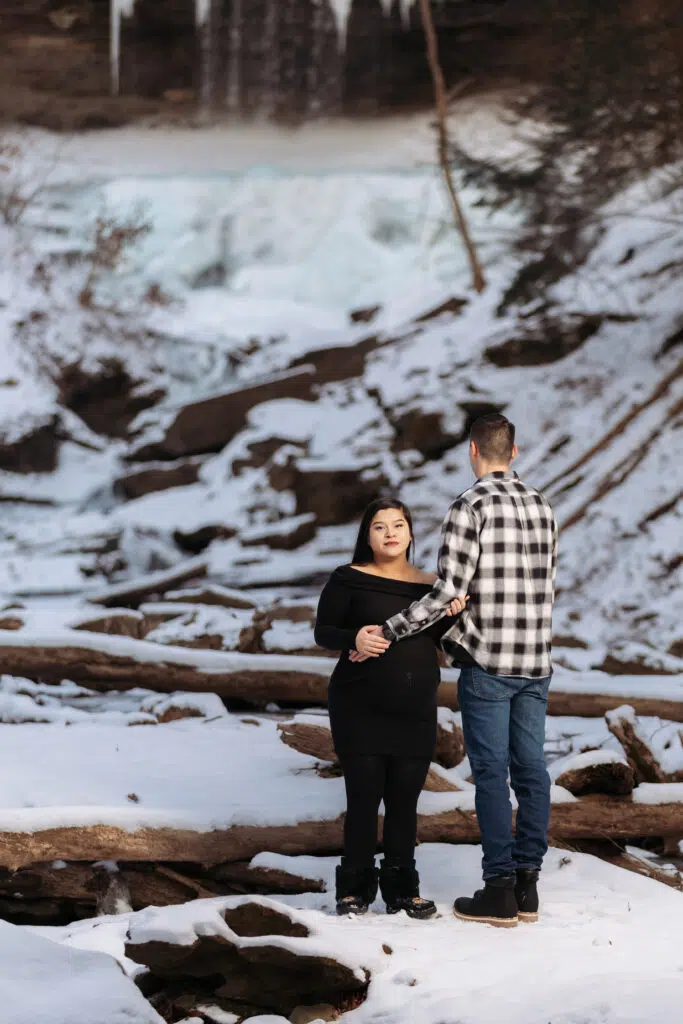 Two people stand on a snowy landscape with a waterfall and icy backdrop, surrounded by logs and winter scenery.