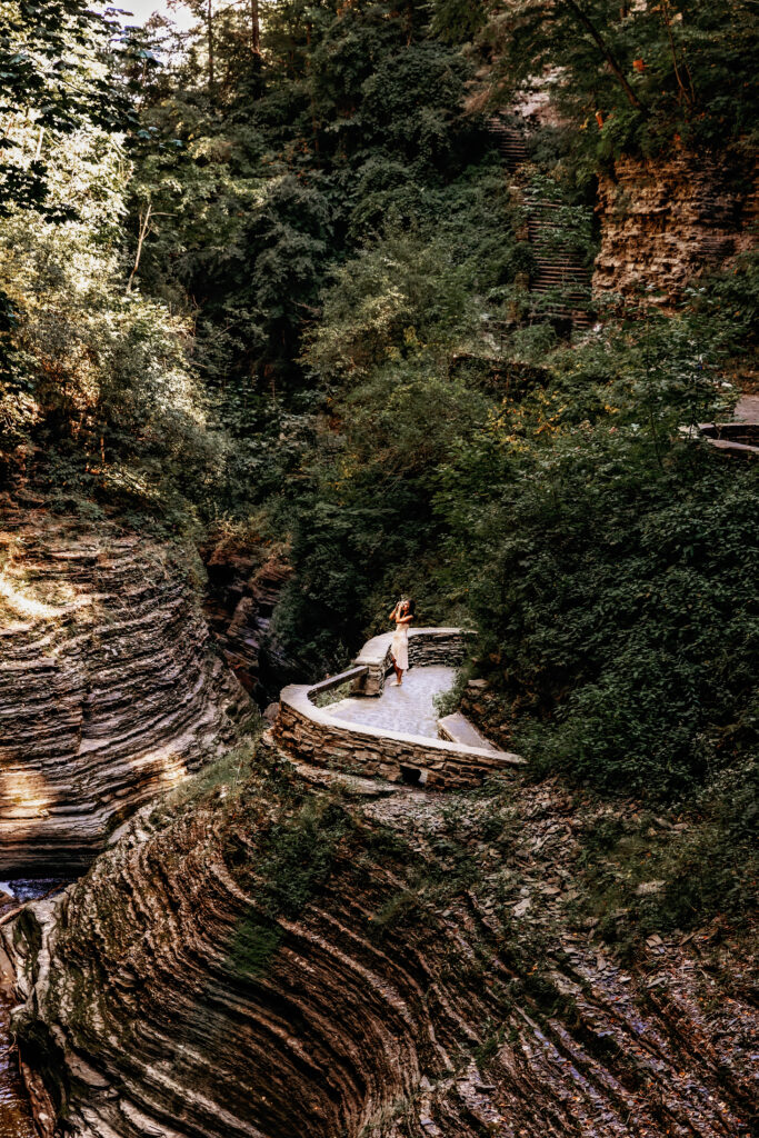 A person stands on a stone path surrounded by lush greenery and layered rock formations at Watkins Glen State Park in New York.