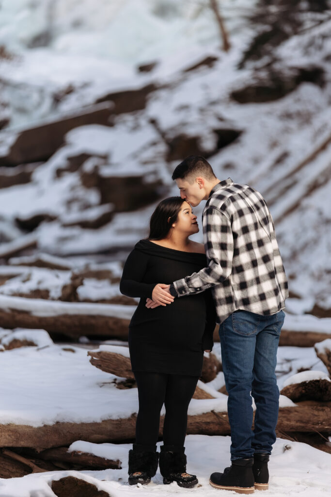 A person kisses another person's forehead in a snowy, rocky landscape, both wearing winter attire, surrounded by logs and snow-covered terrain.