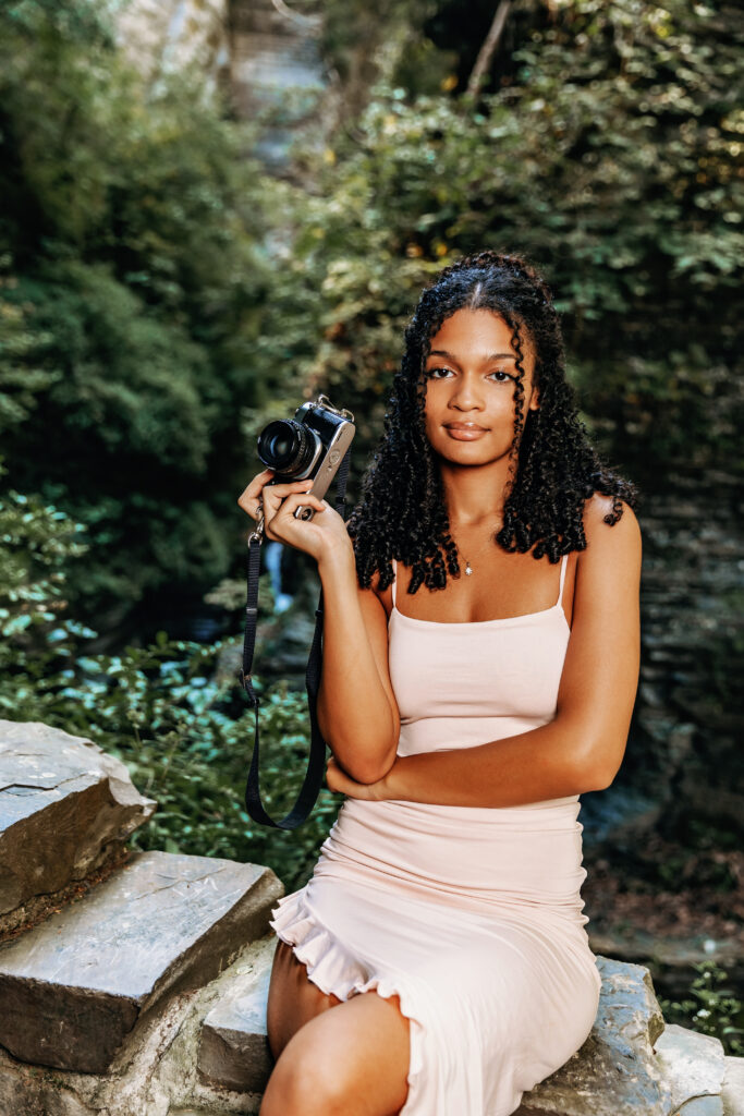 A person in a light dress holds a camera while sitting on stone steps in a lush, green forested area.