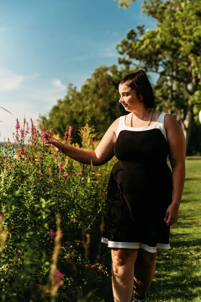A person in a black and white dress admires flowers in a lush, sunlit garden with a backdrop of green trees and a clear sky.