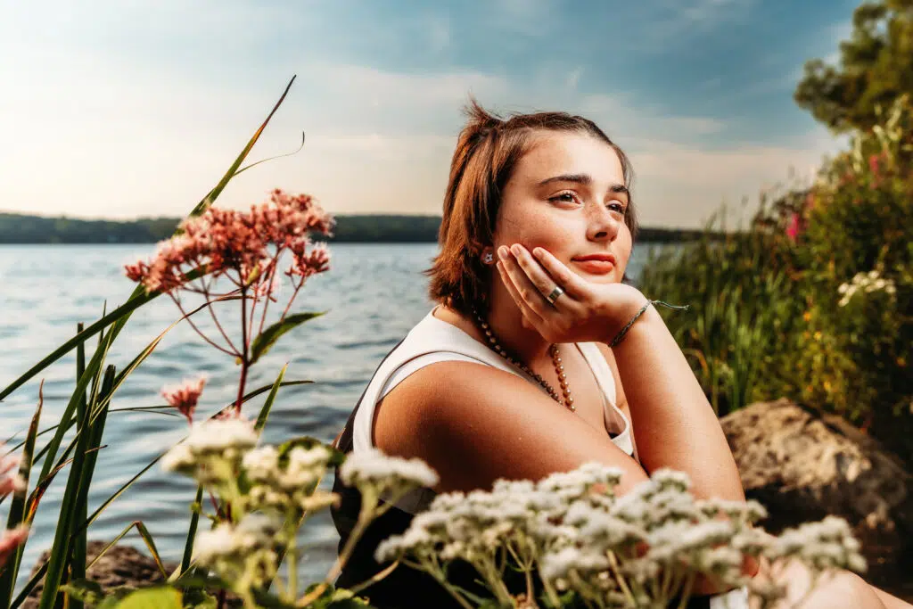 A person sitting by a lake, surrounded by wildflowers and greenery, gazes thoughtfully into the distance under a clear sky.