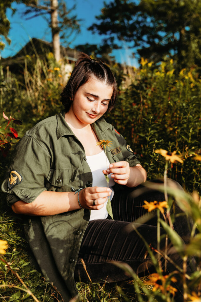 A person sits in a meadow, holding a yellow flower and surrounded by greenery and wildflowers, with trees and a building in the background.