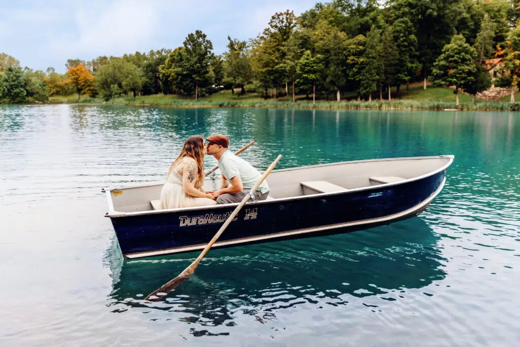 Two people are sitting in a rowboat on a serene lake, surrounded by trees and greenery, sharing a kiss.