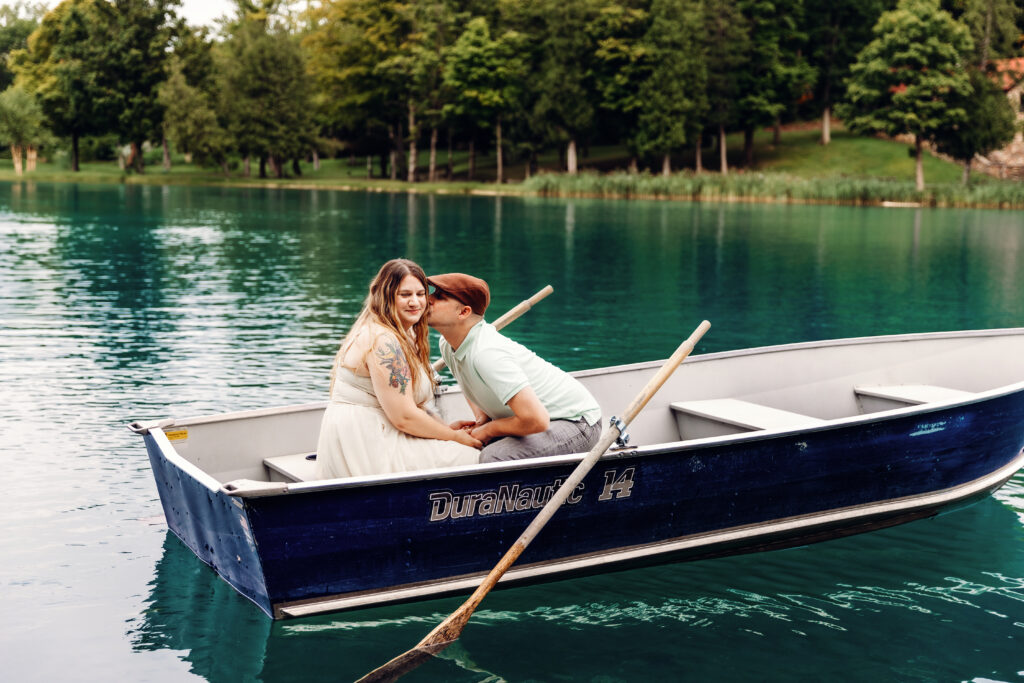 Two people are sitting in a blue rowboat on a serene lake surrounded by lush trees, sharing an intimate moment.