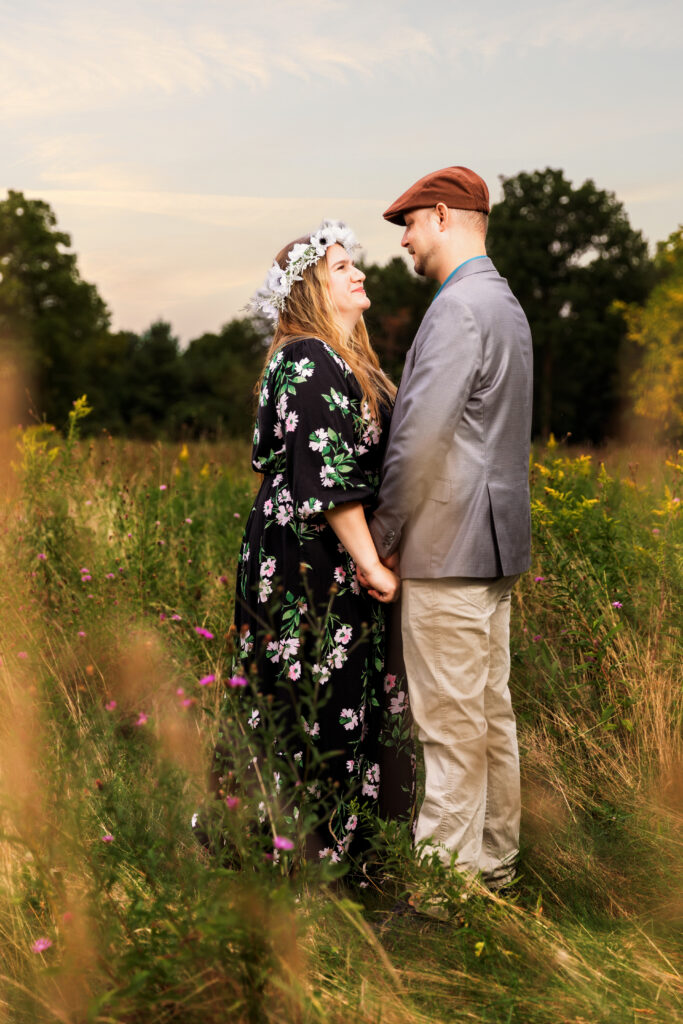 Two people stand in a meadow, holding hands and facing each other, surrounded by tall grass and wildflowers, with trees in the background.