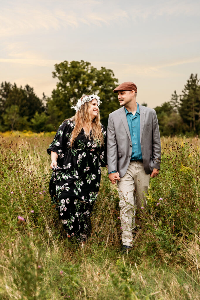 Two people holding hands, walking through a meadow with tall grass and wildflowers, under a partly cloudy sky and surrounded by green trees.
