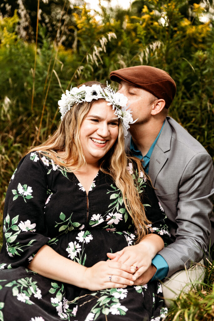Two people sitting in a field, one wears a floral crown, the other kisses their head, both smiling and holding hands.