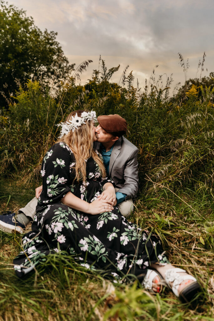 Two people sitting in tall grass kiss at sunset; person wearing floral dress with flower crown, person in blazer with cap.