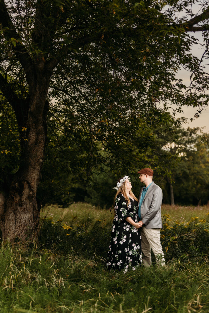 Two people hold hands under a large tree in a lush, green outdoor setting, with one person wearing a floral dress and wreath.