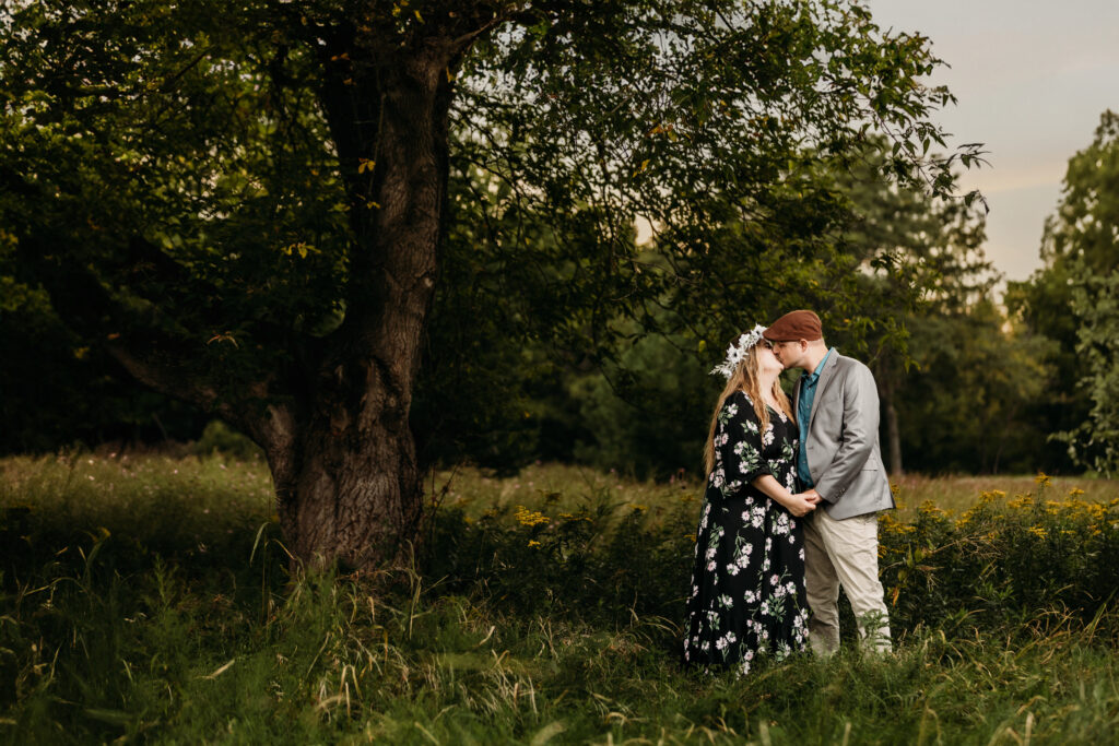Two people share a kiss under a large tree in a lush, green meadow. Flower crown, floral dress, and casual attire.