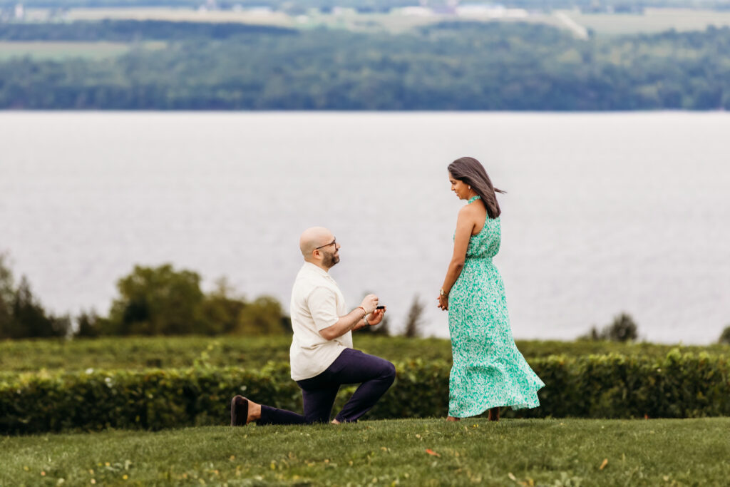 A person proposes on one knee to another in a scenic outdoor setting with a large body of water and hills in the background.