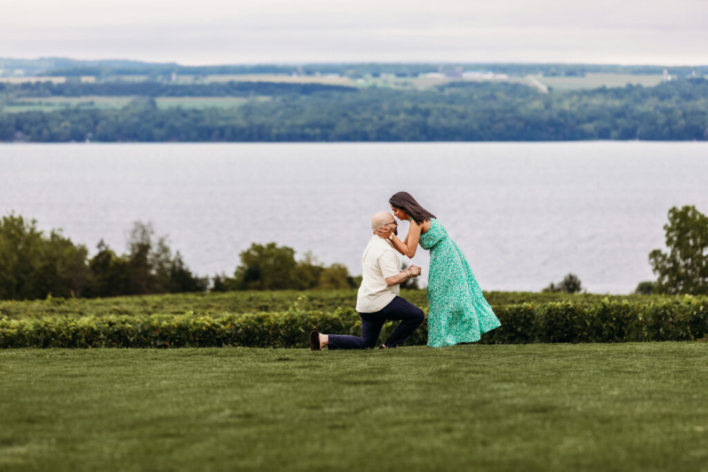A person kneels proposing to another person in a green dress on a grassy lawn by a scenic lake with distant wooded hills.