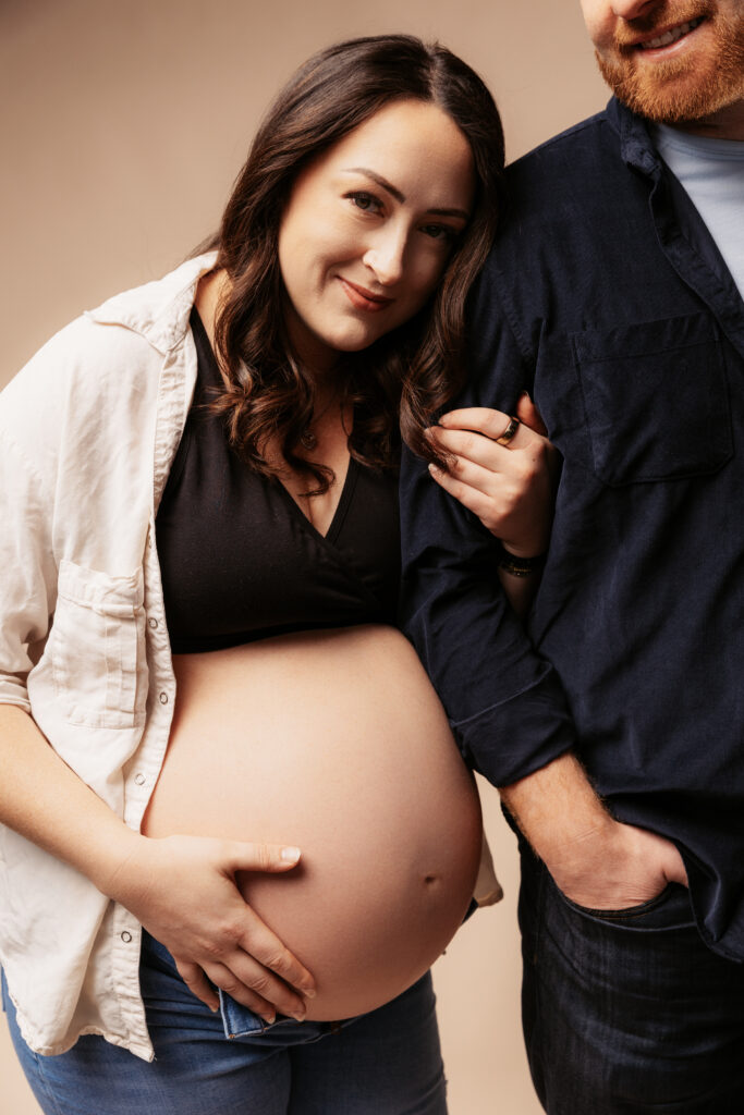 Two people standing closely, one pregnant, smiling gently. Casual attire with a neutral backdrop. Hand placed lovingly on belly.