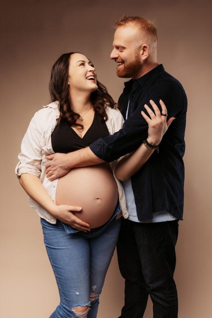 A smiling couple embraces, highlighting the pregnant person's belly. Casual clothing, warm expressions, neutral background suggest a joyful, intimate moment.