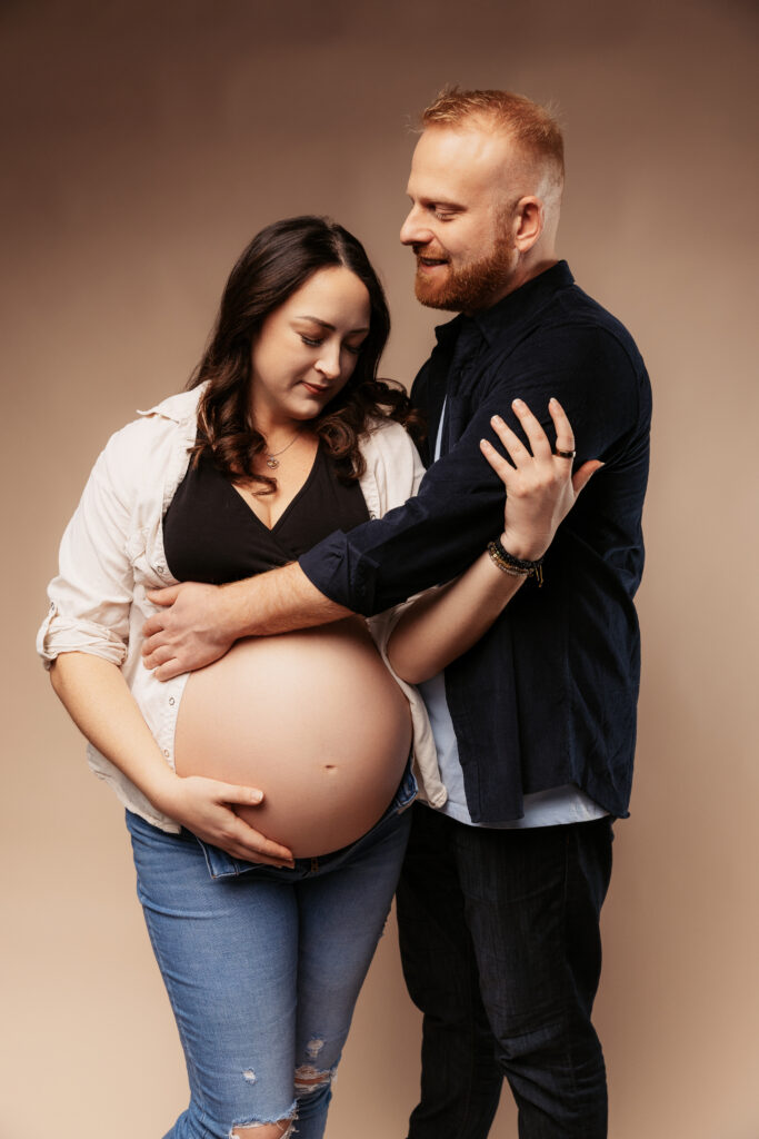 Two people embracing, with one displaying a pregnant belly. They are standing against a plain, neutral background, looking at each other affectionately.