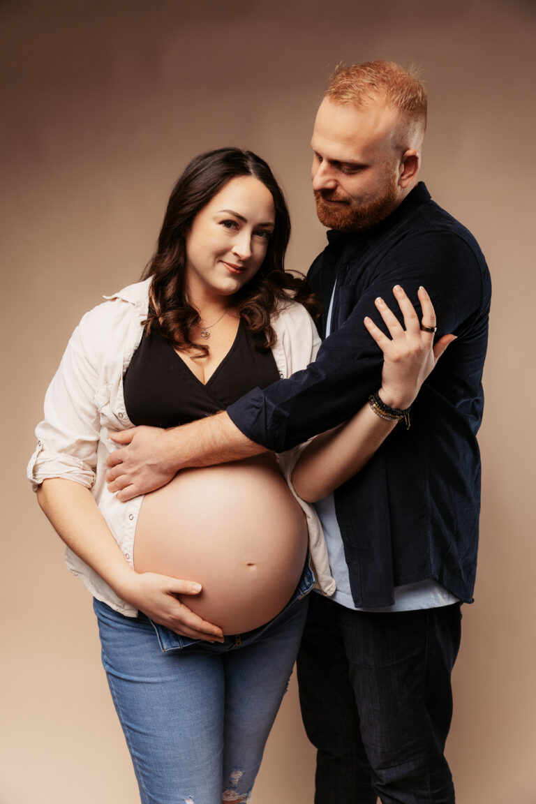 Two people stand closely, one pregnant, embracing affectionately. They wear casual clothes against a neutral background, conveying warmth and anticipation.