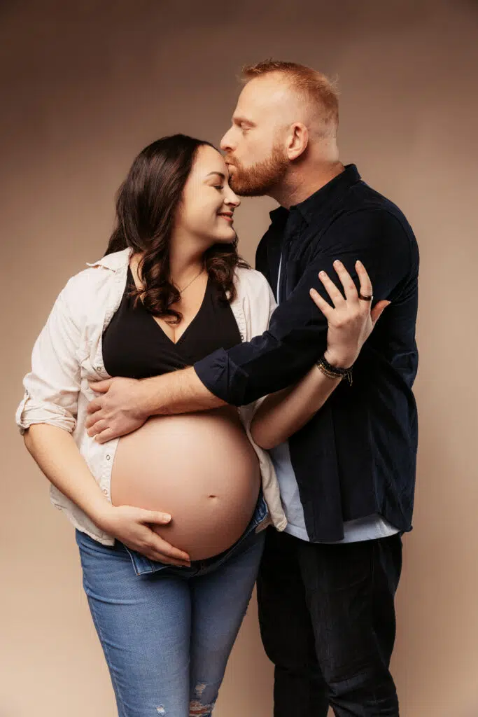 A person kisses another's forehead, embracing a baby bump. Both are smiling, dressed casually, against a neutral background, conveying warmth and affection.