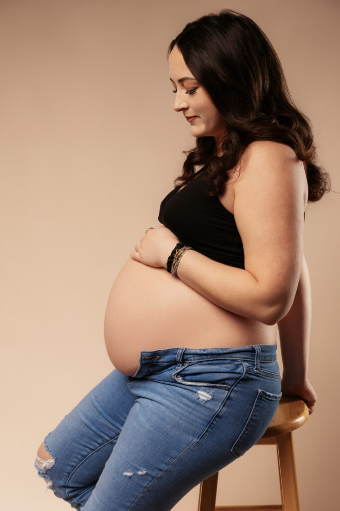 A person with long hair sits on a stool, wearing a black top and ripped jeans, cradling a pregnant belly against a plain background.