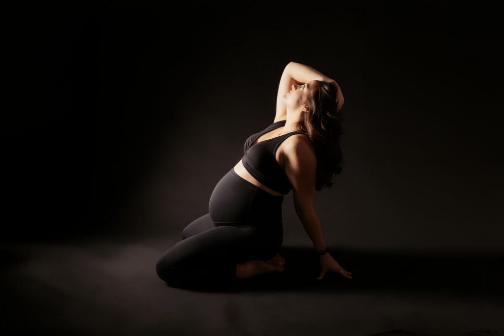 A person in black attire poses dramatically on the floor against a dark background, conveying strength and elegance in maternity.