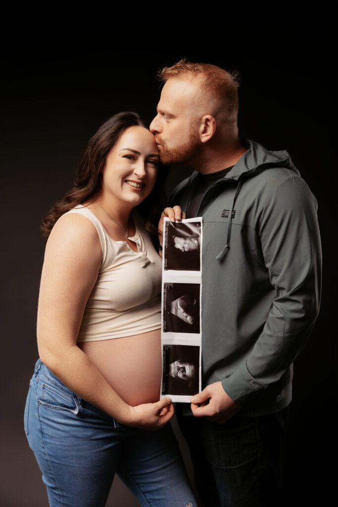 A person kisses another's head while they display ultrasound images, showing a pregnancy. The setting is studio-lit with a dark background.