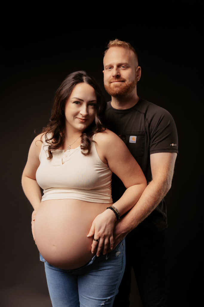 Two people pose together in a studio setting. The foreground person is visibly pregnant, both smiling warmly against a dark background.