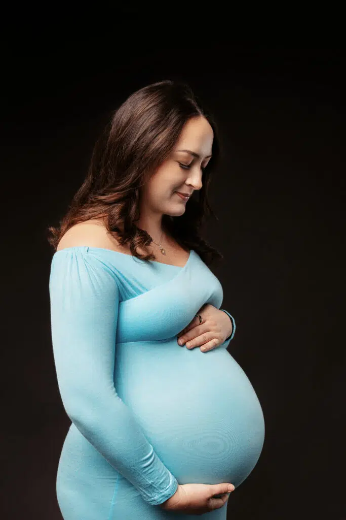 A person in a blue dress, pregnant and smiling gently, poses against a dark background, displaying a calm and reflective demeanor.