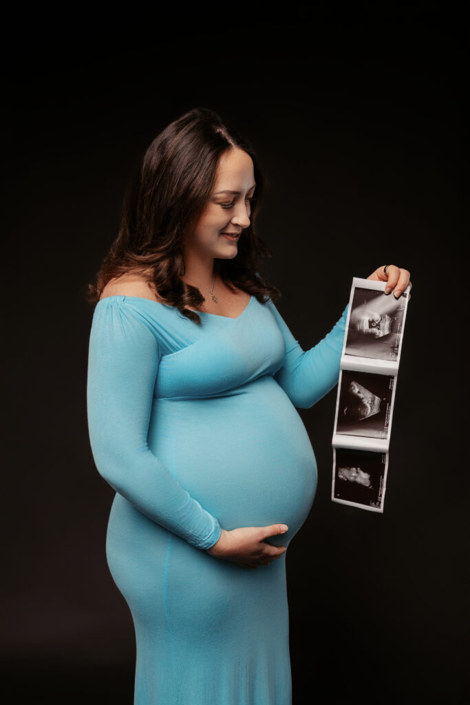 A smiling person in a blue dress holds ultrasound images, cradling their belly, set against a dark background.