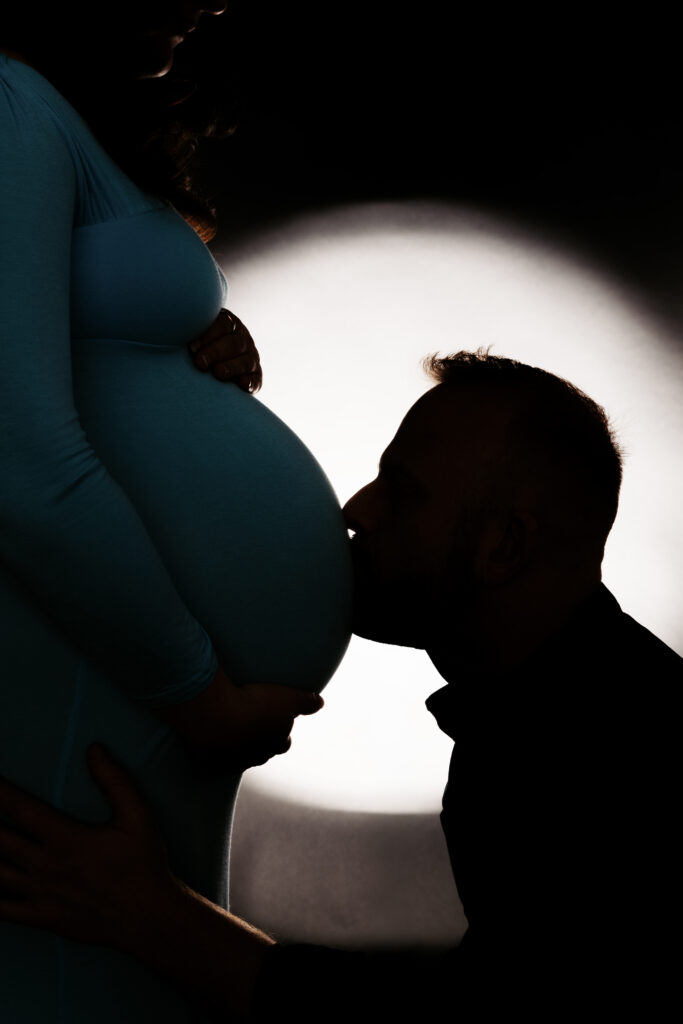 Silhouette of a person kissing another person's pregnant belly, set against a contrasting soft light backdrop. Tender and intimate moment captured.