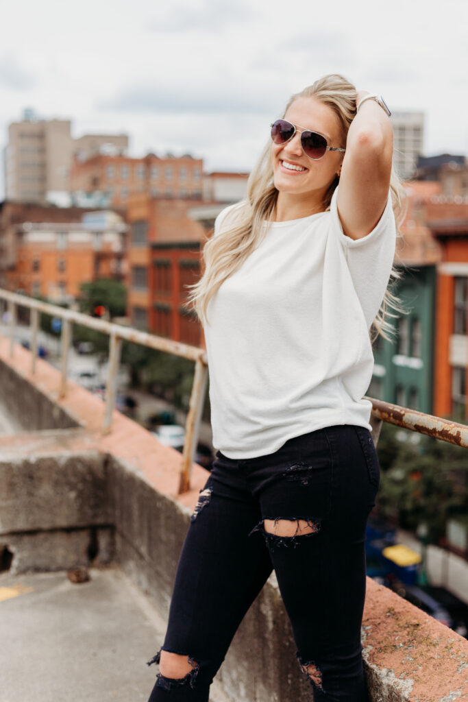 A person stands smiling on an urban rooftop, wearing sunglasses, a casual white top, and ripped black jeans, with a cityscape backdrop.