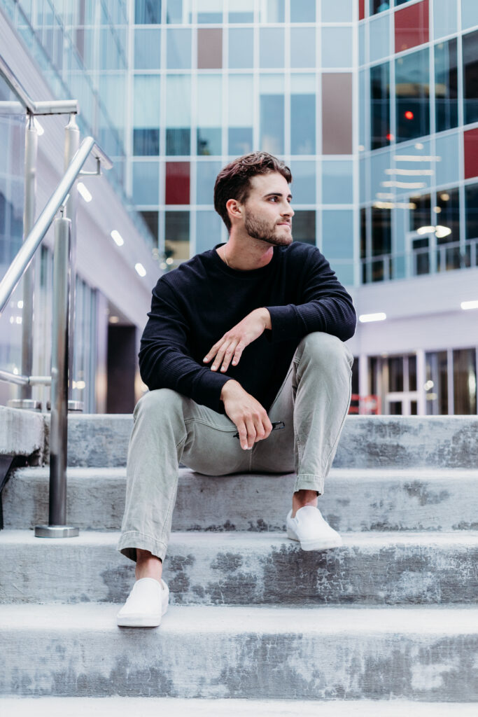 A person sits on concrete steps, looking to the side, with a reflective glass building backdrop. They're wearing a dark sweater, light pants, and white shoes.