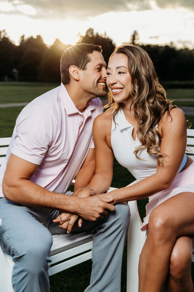 Two people are seated closely together outdoors, smiling and holding hands. The background features a sunlit sky and dark silhouetted trees.