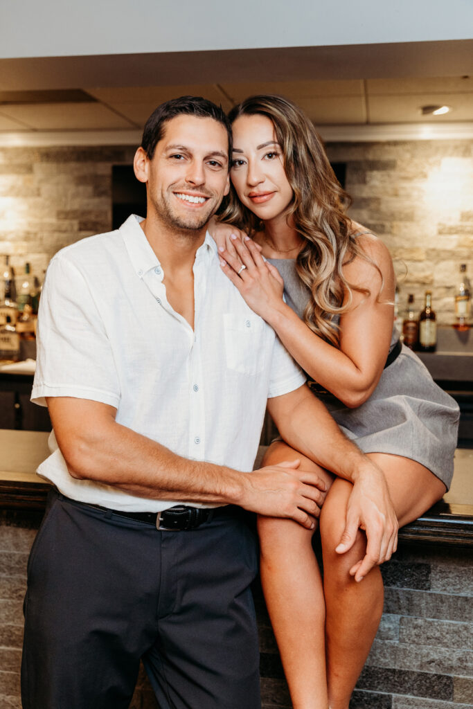 Two people posing affectionately in a bar or restaurant setting, with a brick wall and bottles in the background.