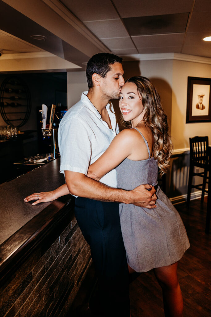 A person kisses another on the forehead, embracing near a bar counter, with a framed photo on the wall in the background.
