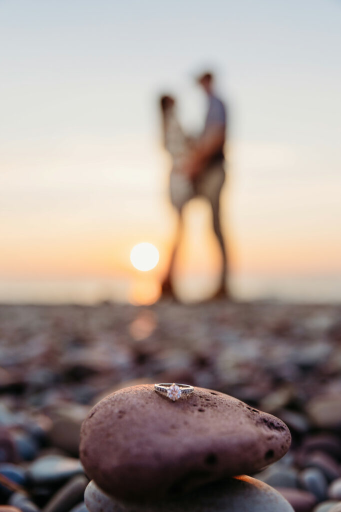 An engagement ring placed on a stone, sharply focused, with a blurry background featuring two people standing by the sea at sunset.
