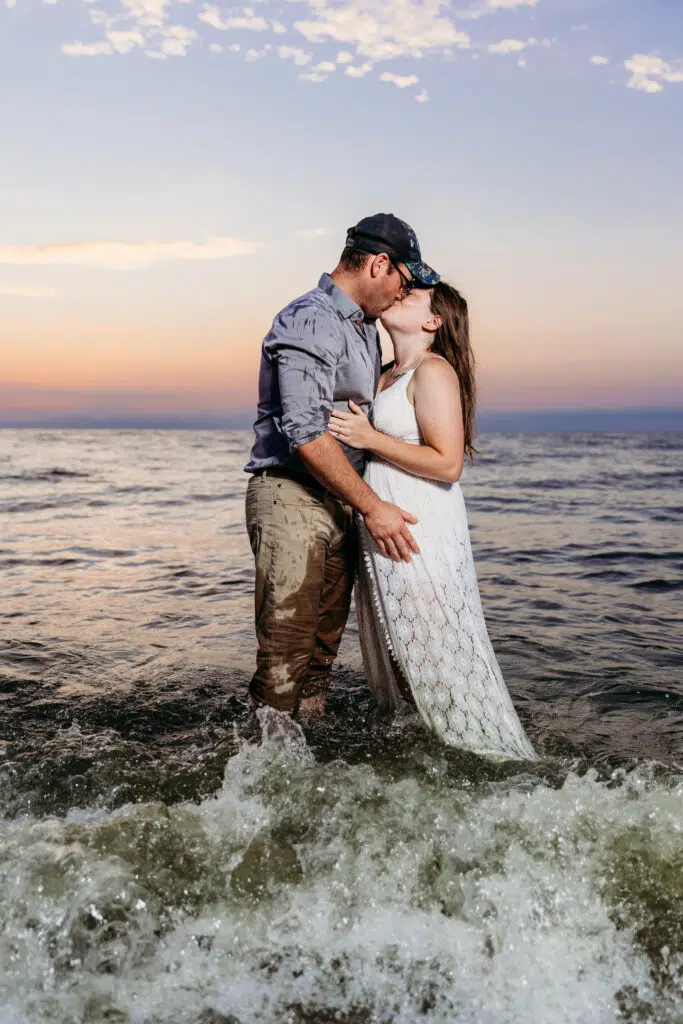 Two people are kissing in shallow water against a sunset sky. They're embracing, waves around them, creating a romantic and serene atmosphere.