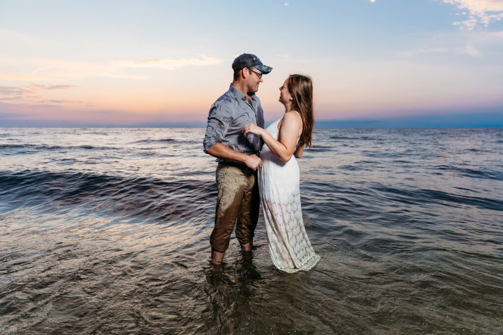 Two people standing in the sea holding hands, looking at each other lovingly, during sunset with a colorful sky in the background.
