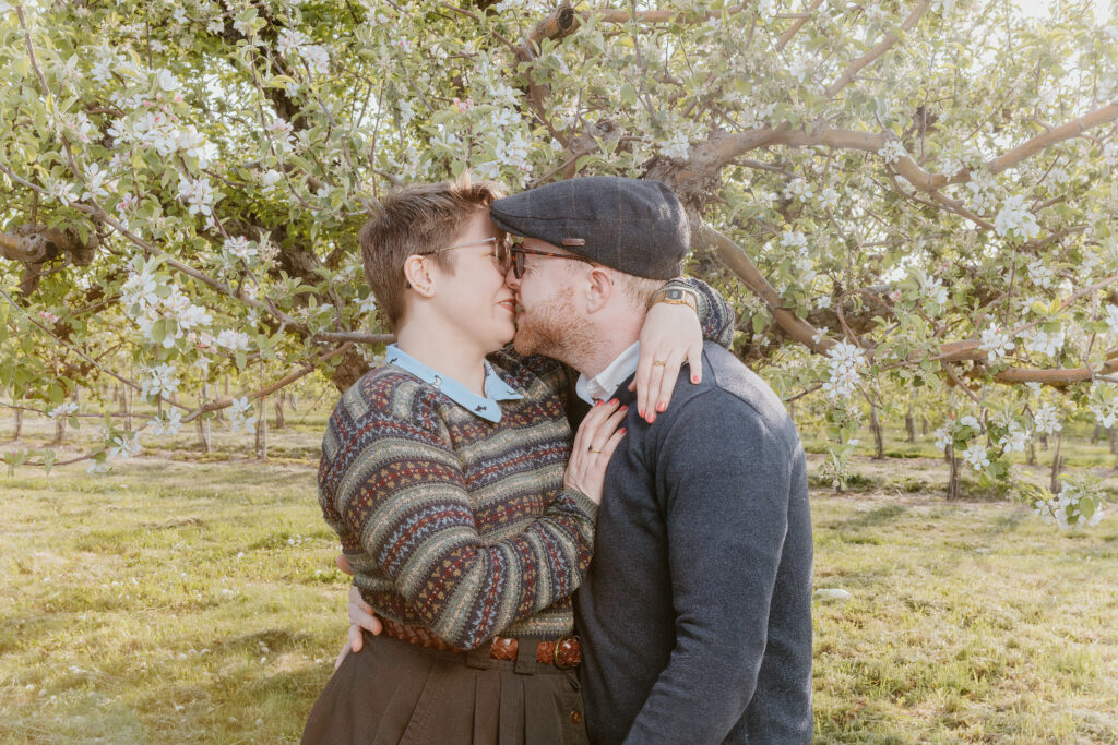 Two people are sharing a romantic moment, kissing among blossoming trees. They're dressed in cozy, casual clothes with a vintage feel, immersed in a serene orchard.
