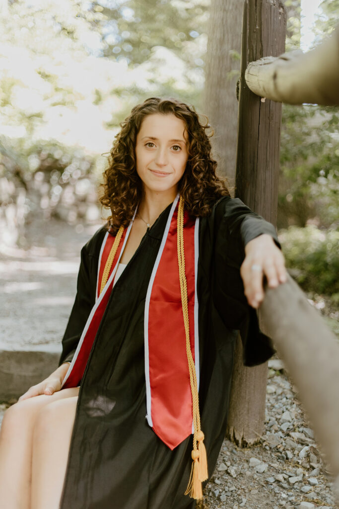 A person in a black graduation gown with red accents smiles gently, leaning against a wooden post amidst a dappled sunlight background in a wooded area.
