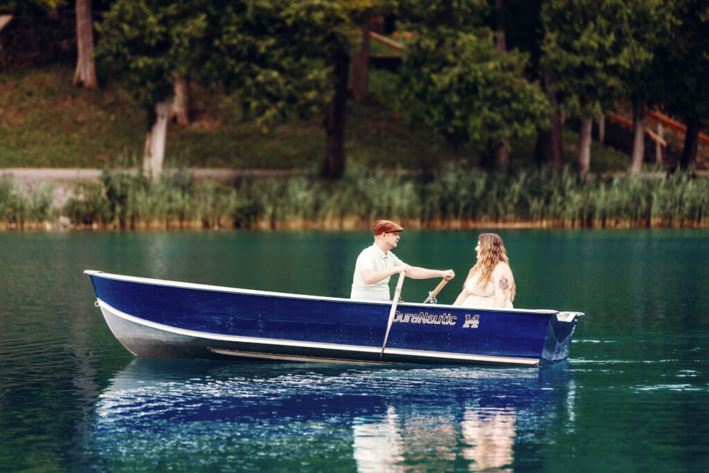 A person rows a blue boat with another person on a calm lake, surrounded by lush greenery and trees in the background.
