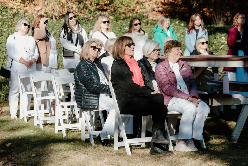 A group of people seated outdoors on white chairs, attentively watching an event in a sunny garden setting with surrounding greenery.