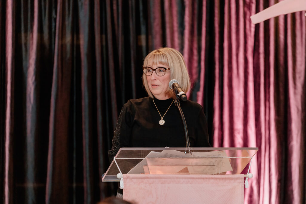 A person with glasses speaks at a podium with a microphone, against a backdrop of dark, textured curtains.