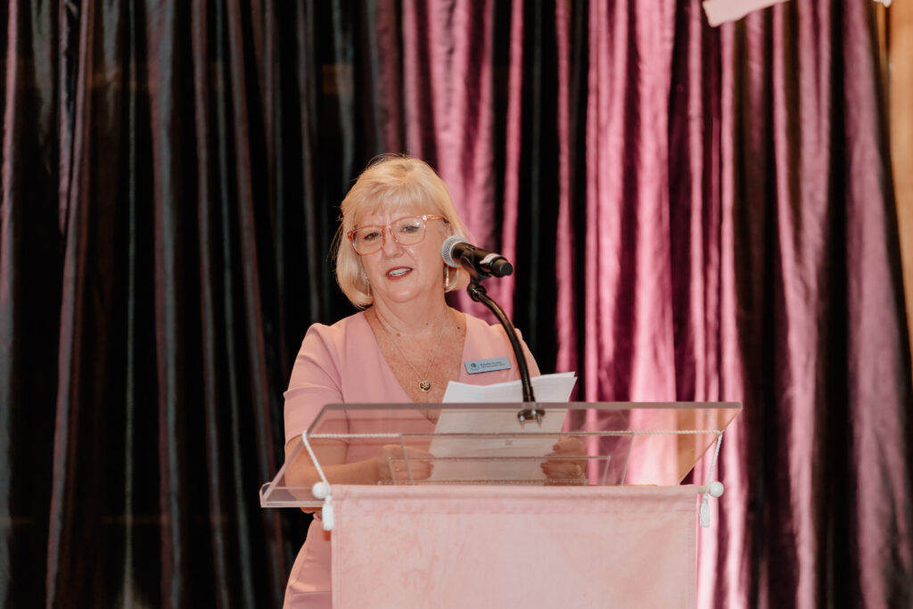 A person in pink speaks at a podium with a microphone, against a dark, shimmering curtain backdrop.