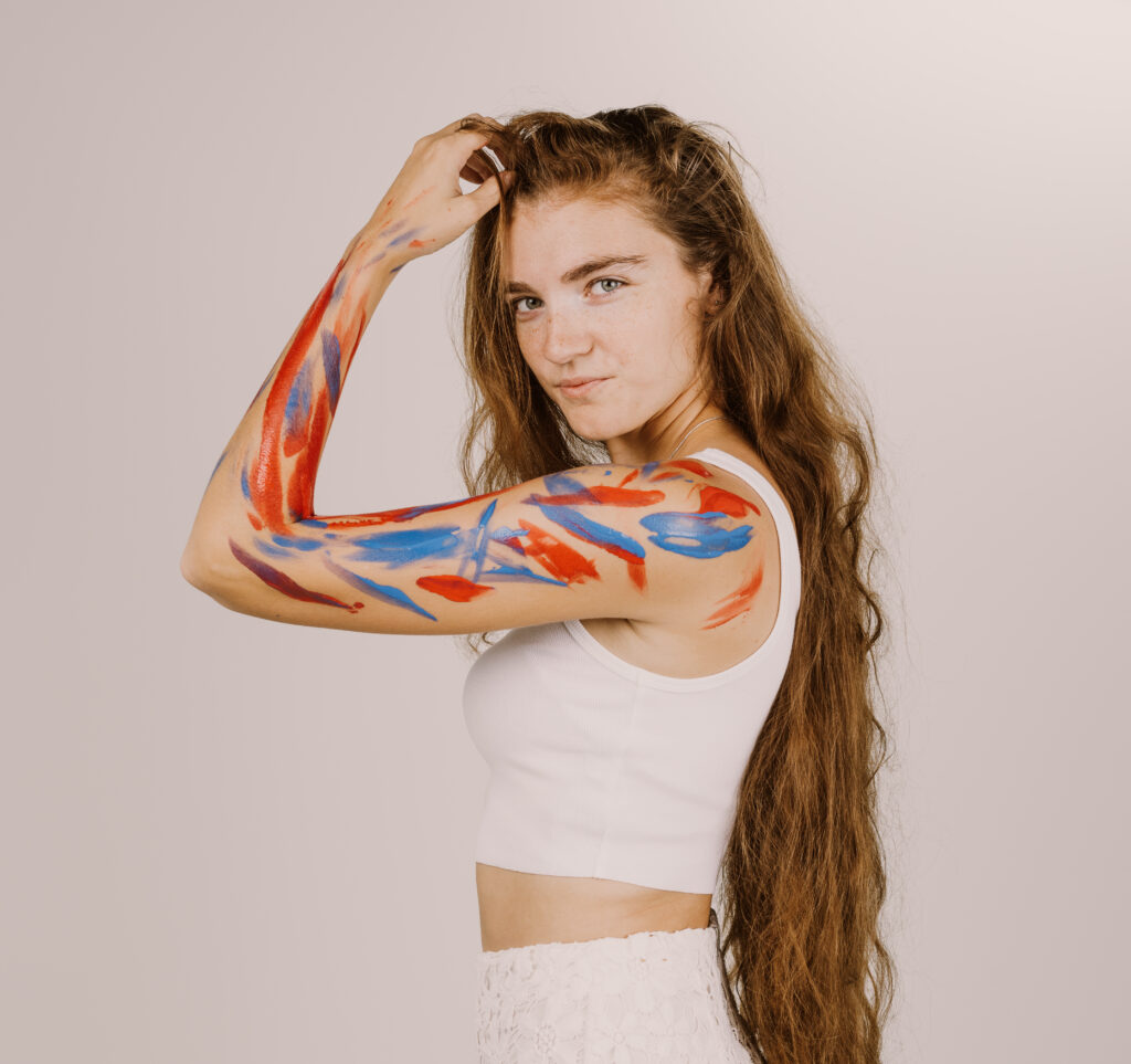 A person with long hair stands against a plain background, showcasing an arm painted with vibrant blue and red strokes, giving a confident look.