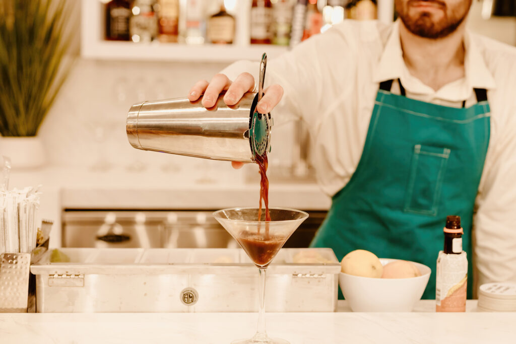 A person in an apron pours a cocktail into a glass at a bar, with bar tools and ingredients visible in the background.