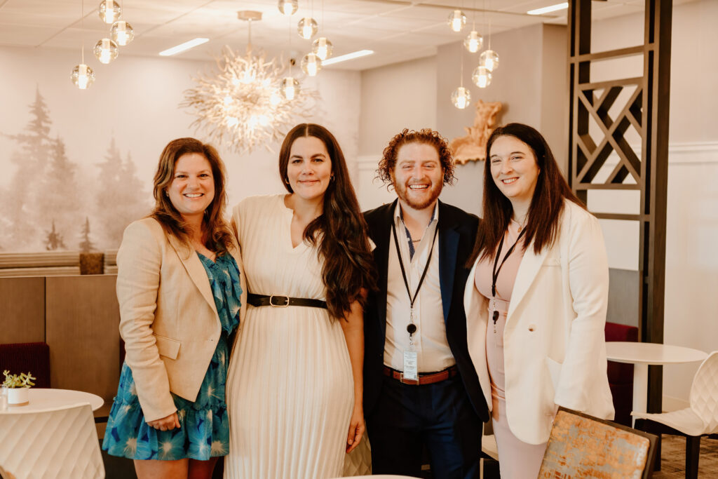 Four smiling people are standing together indoors with modern decor, featuring pendant lights and artistic wall design.